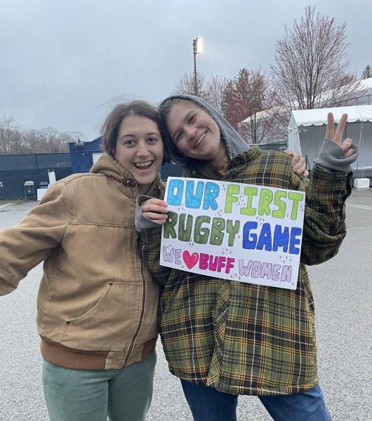 Sam Espach and Effie Hossfeld of the scarlet holding a poster that reads “Our first rugby game. We (heart) buff women.”